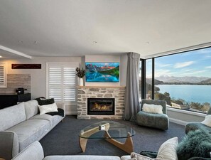 A modern living room with a fireplace, a large flat-screen TV, and a view of a lake and mountains. - Above the Lake | Queenstown Sanctuary (Queenstown)