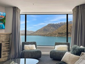 Ein Zimmer mit großem Fenster und Blick auf die Berge, zwei Sessel mit flauschigen Kissen, ein Glastisch und ein Gemälde an der Wand.