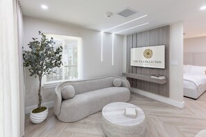 A modern hotel room with a sofa, a round coffee table, a potted plant, a mounted TV, and a bed.
