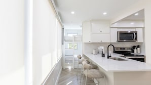 Cocina moderna con armarios blancos, microondas de acero inoxidable y horno empotrado. Hay tres taburetes de bar y una ventana que proporciona luz natural.