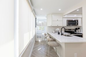 A modern kitchen with white cabinetry, a stainless steel microwave, and a built-in oven. There are three bar stools and a window providing natural light. - Octa Collection on Front Beach (Panama City Beach)