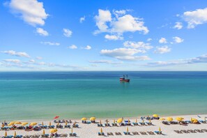 A beach with clear blue water, a boat in the distance, and rows of sun loungers with umbrellas.