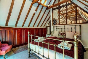 Ein Schlafzimmer mit schrägverdeckter Decke, einem Bett mit Metallgestell, Holzschränken und einem rot gepolsterten Sessel.