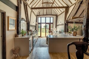 Une cuisine rustique dotée d’un grand îlot central, de poutres en bois et offrant une vue sur l’extérieur par la porte.