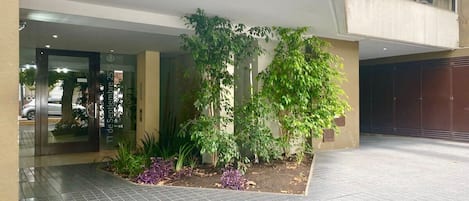 Une entrée d’immeuble avec une zone pavée, un petit jardin agrémenté de plantes et une porte d’entrée en verre.