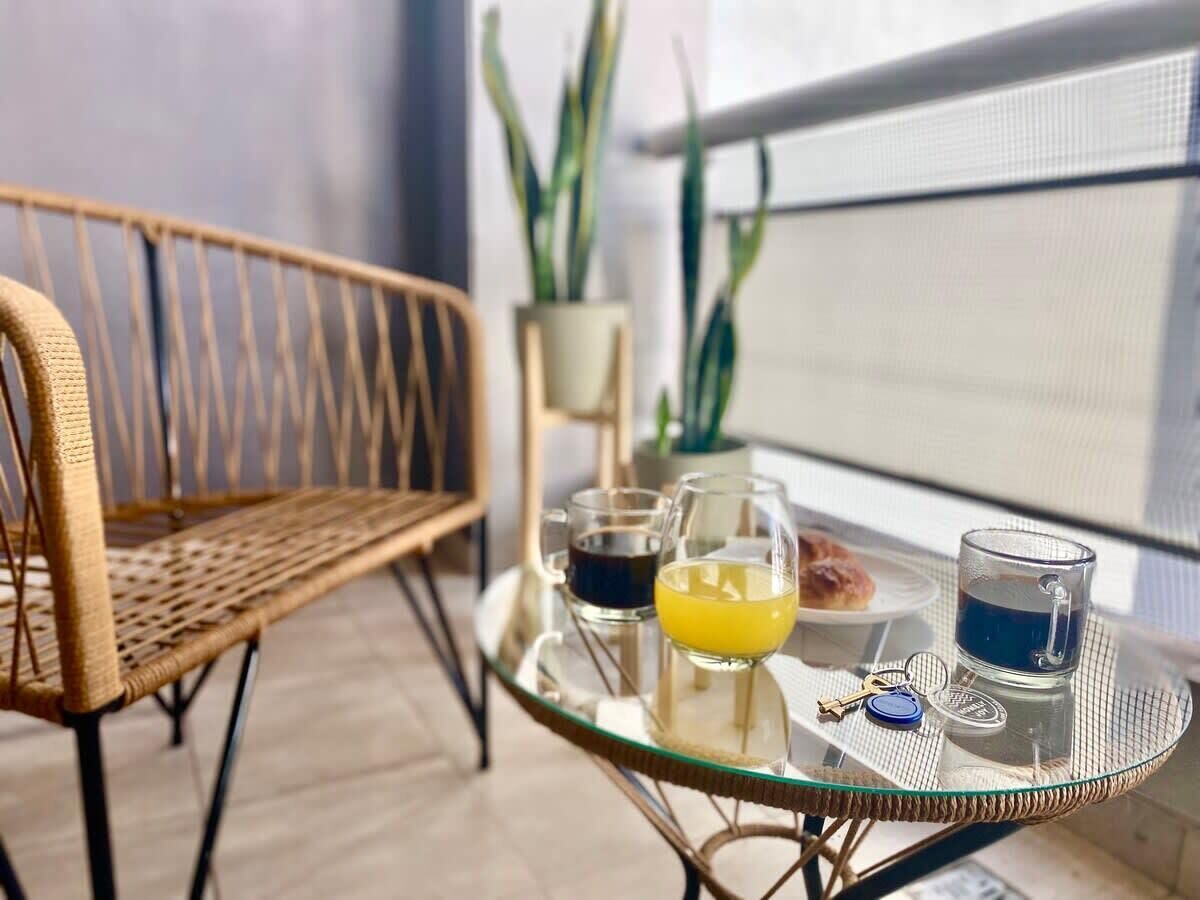 A glass table with drinks and keys, a wicker chair, and a potted plant in a room with a view.