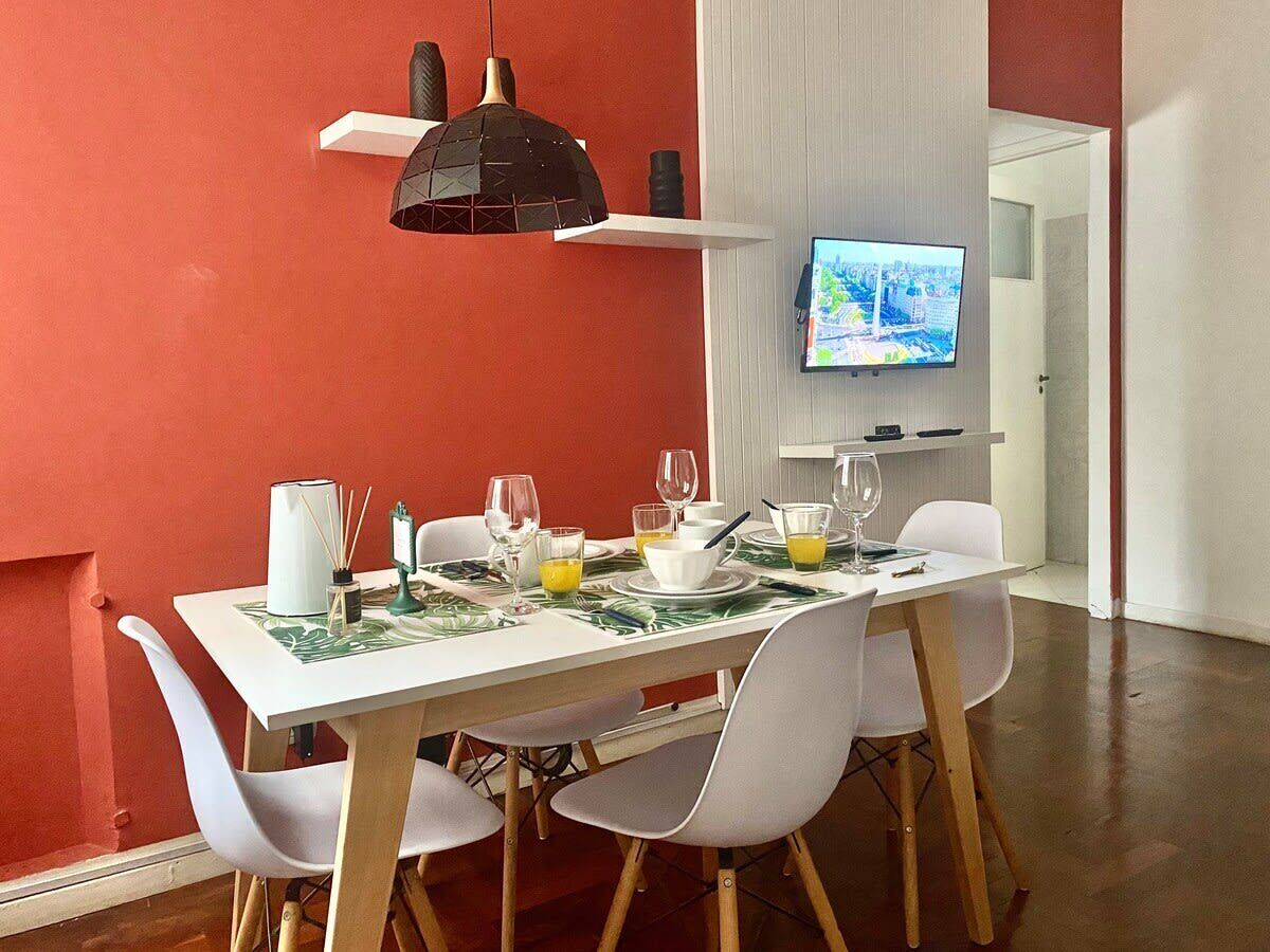 Un coin repas avec une table dressée pour quatre personnes, des chaises blanches, un luminaire suspendu noir et un téléviseur fixé au mur.