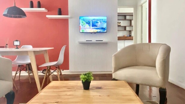 A modern interior with a wooden coffee table, a beige armchair, a television mounted on a white wall, and a dining area with chairs and a table. - 157- Apt. with Great Location for 3 p. in Recoleta (Buenos Aires)