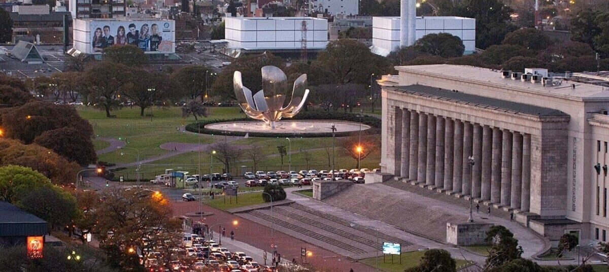 A large building with a classical architectural style, a central open space with a unique sculpture, and a parking area with multiple cars.