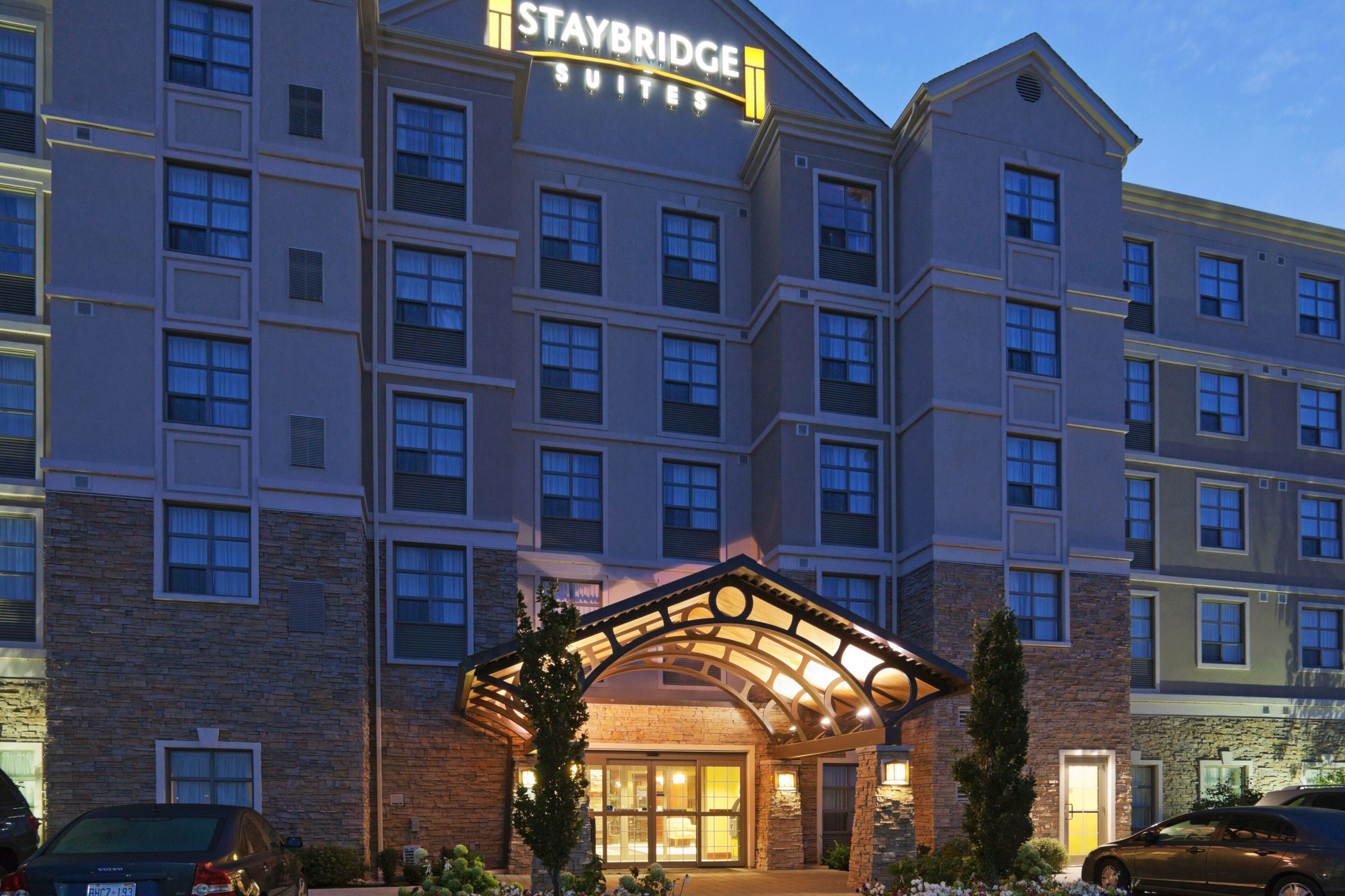 Un hotel de varios pisos con un llamativo letrero de Staybridge Suites, fachada de piedra y entrada cubierta con techo curvo.