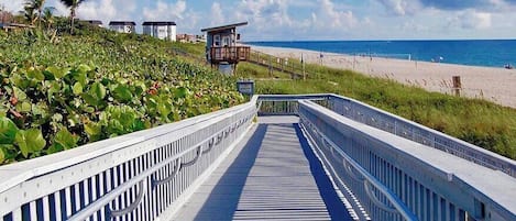 A boardwalk leading to a beach with a lifeguard tower, vegetation, and a clear sky.