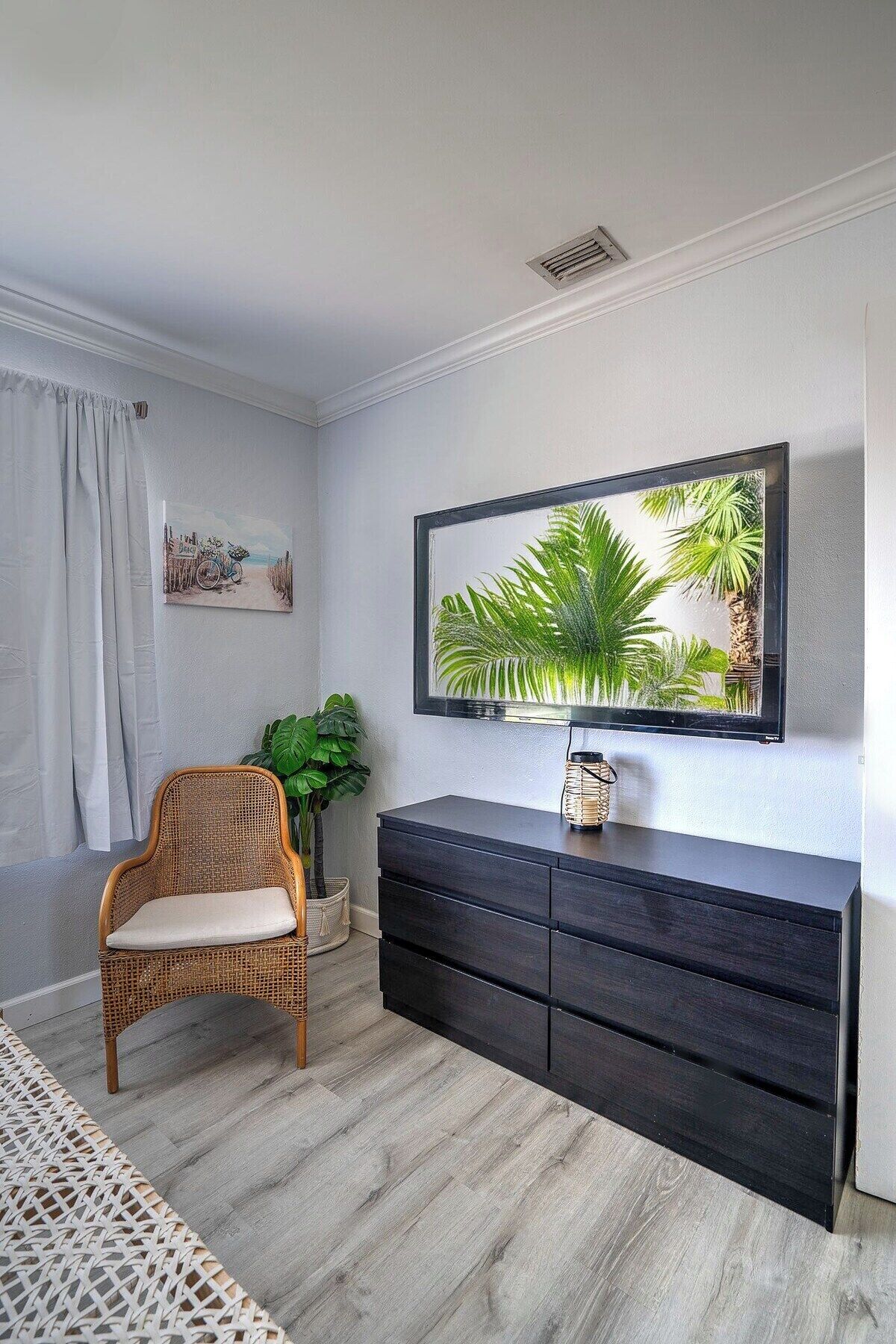 A room with a wicker chair, a dark wooden dresser, a framed picture, and a large mirror with a tropical scene.