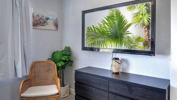 A room with a wicker chair, a dark wooden dresser, a framed picture, and a large mirror with a tropical scene.