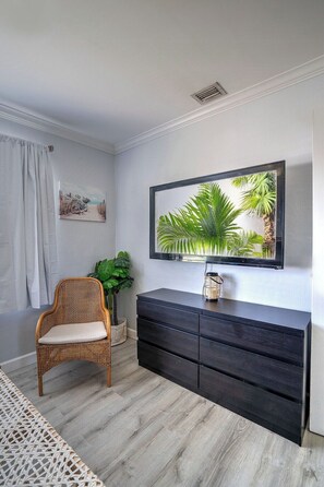 A room with a wicker chair, a dark wooden dresser, a framed picture, and a large mirror with a tropical scene. - Renovated Coastal Hideaway • 2BR • Minutes to Beach • Private Parking & 65” TV (Boynton Beach)