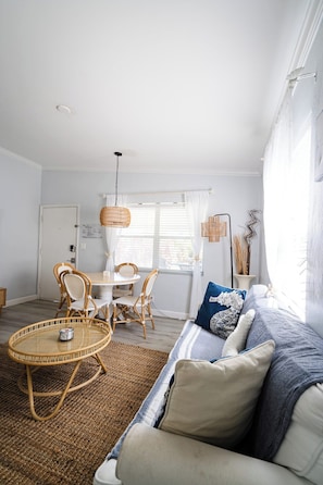 A cozy living room with a round wicker coffee table, a sofa with patterned cushions, and a dining area with wicker chairs.