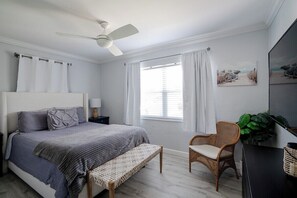 Ein Schlafzimmer mit einem Bett, einem Nachttisch, einem Sessel, einem Fenster mit Vorhängen, einem Deckenventilator und einem an der Wand befestigten Fernseher.