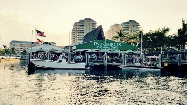 A waterfront restaurant with a green awning labeled 'TWO GEORGES Waterfront Grille.' Boats are docked in the water, and there are buildings in the background. - Renovated Coastal Hideaway • 2BR • Minutes to Beach • Private Parking & 65” TV (Boynton Beach)