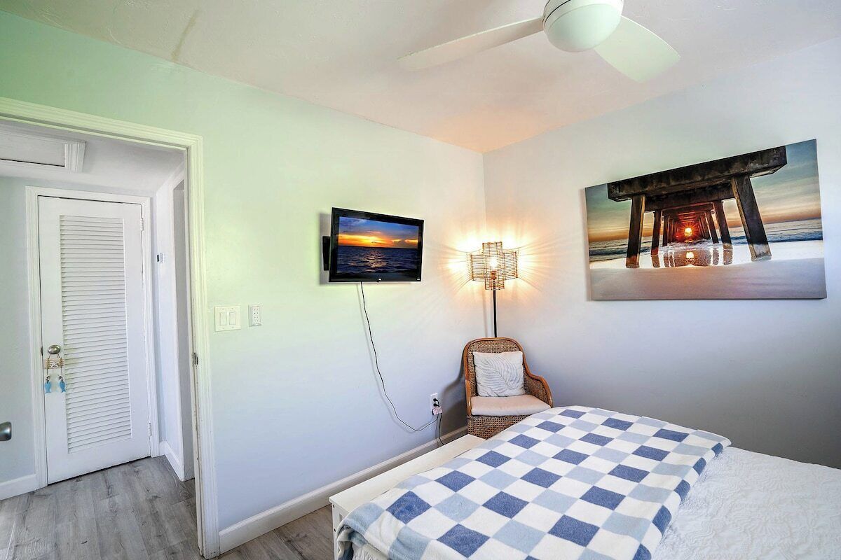 A room with a bed, a television mounted on the wall, a chair, and a painting of a pier.