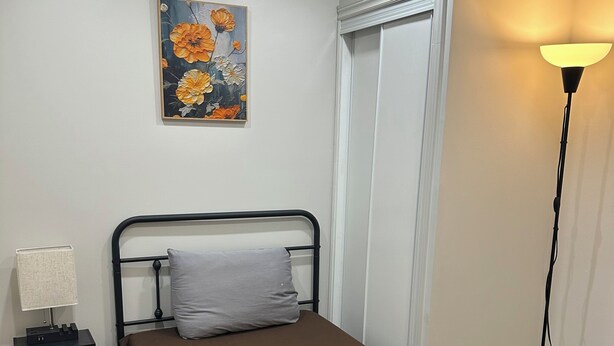 A single bed with a brown mattress, a grey pillow, and a white towel on top. There is a nightstand with a lamp and a tissue box. A painting of flowers hangs on the wall.