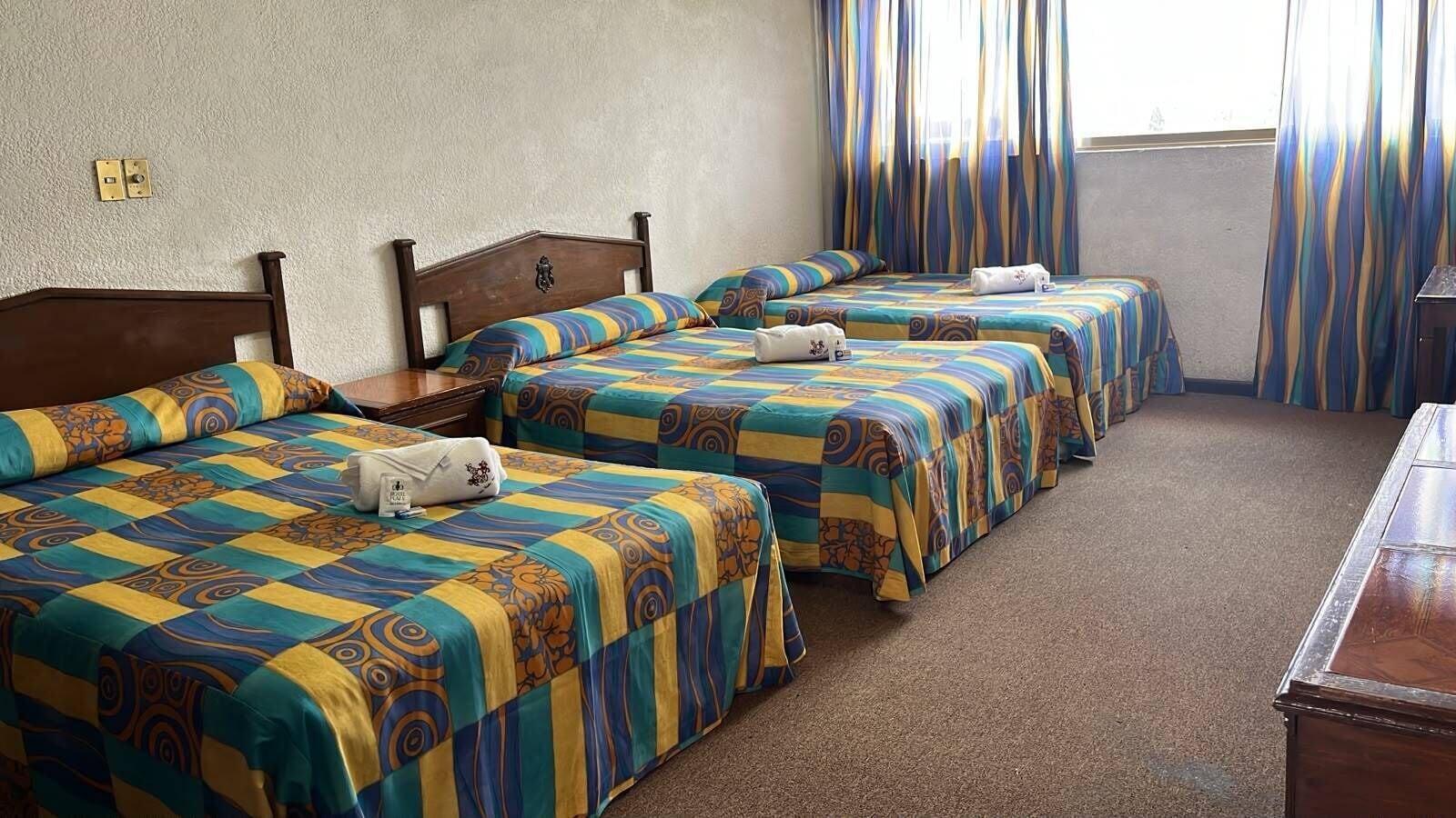 Habitación de hotel con tres camas, cada una con una colcha y almohadas estampadas. Se observa un aparador de madera y una ventana con cortinas.