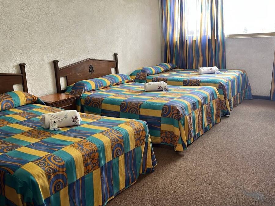 A hotel room with three beds, each with a patterned bedspread and pillows. There is a wooden dresser and a window with curtains.