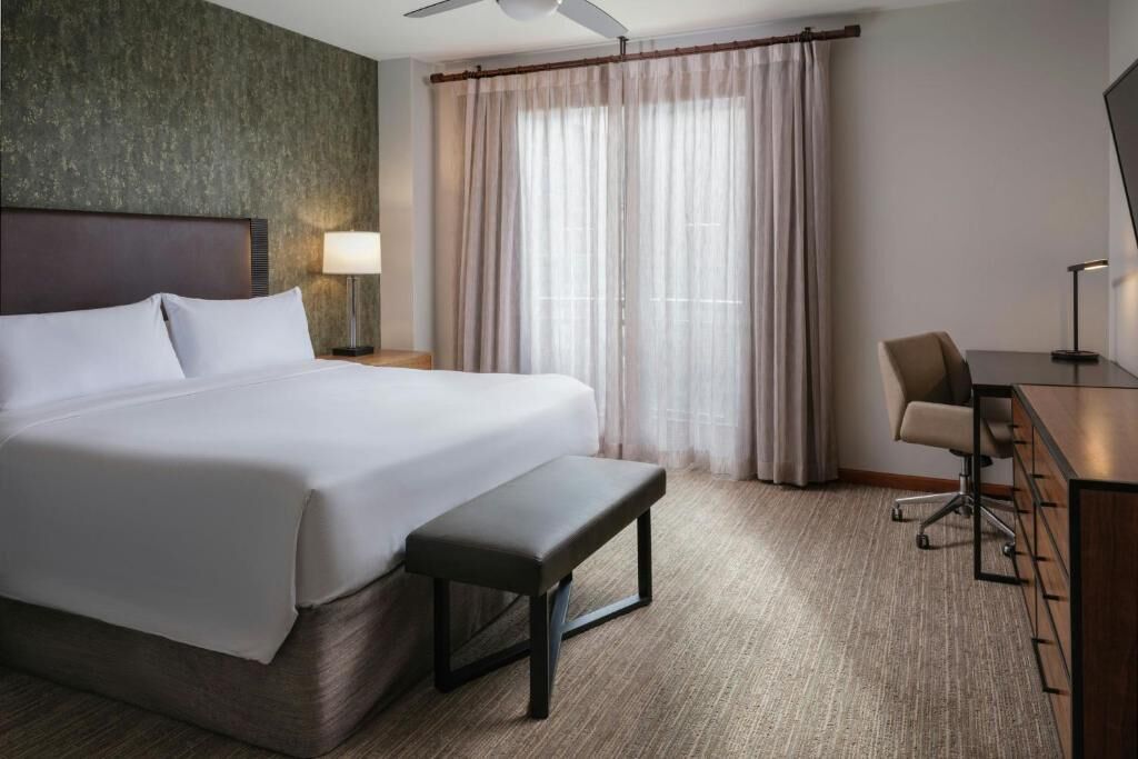 A hotel room with a large bed, a desk, a chair, a bench, a lamp, and a window with curtains.