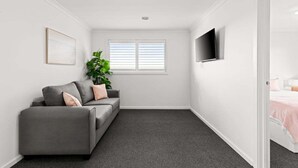 A room with a grey sofa, a potted plant, a flat-screen TV, and a window with blinds. - Scentbark Family Retreat (Surf Coast)