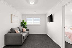 A room with a grey sofa, a potted plant, a flat-screen TV, and a window with blinds. - Scentbark Family Retreat (Surf Coast)
