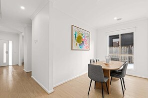 A dining area with a wooden table and chairs, a painting on the wall, and a window with a view. - Scentbark Family Retreat (Surf Coast)