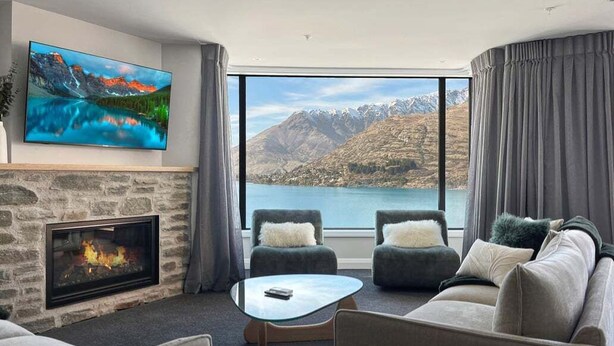 A modern living room with a stone fireplace, a large flat-screen TV, a sofa, two armchairs, and a view of mountains and a lake. - Above the Lake | Lakeview Getaway (Queenstown)