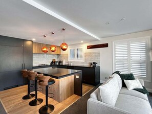 Eine moderne Küche mit Kochinsel, Barhocker, Pendelleuchten und Blick in einen Wohnbereich mit einer Couch.