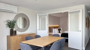 A modern room with a wooden dining table, grey upholstered chairs, a round mirror, and a bed with a white headboard. - Above the Lake | Lakeview Getaway (Queenstown)