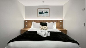 A hotel room with a neatly made bed, a headboard with a picture, and a bedside table with a lamp. - Above the Lake | Lakeview Getaway (Queenstown)