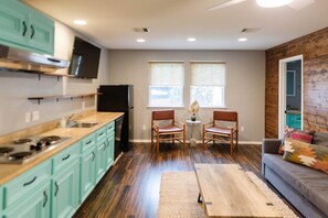 A compact kitchen with teal cabinets, a black refrigerator, and a stove. There are two chairs and a small table with a vase. The room has wooden flooring and a gray wall with two windows. - Chef's Kitchen · Game Room+Casita · Group Ready! (Austin)
