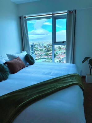 A hotel room with a large bed, a view of a cityscape through a window, and a potted plant. - Relaxing, Cozy Apartment on the 20th Floor! AC Wi-Fi Pool (San Jose)