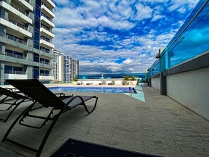A pool area with lounge chairs, a clear view of the ocean, and a high-rise building in the background. - Relaxing, Cozy Apartment on the 20th Floor! AC Wi-Fi Pool (San Jose)