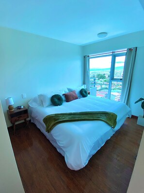 A hotel room with a large bed, a bedside table with a lamp, and a view of a cityscape through a large window. - Relaxing, Cozy Apartment on the 20th Floor! AC Wi-Fi Pool (San Jose)