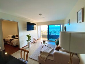 A modern living room with a sofa, a coffee table, and a TV mounted on the wall. There is a balcony with a city view. - Relaxing, Cozy Apartment on the 20th Floor! AC Wi-Fi Pool (San Jose)