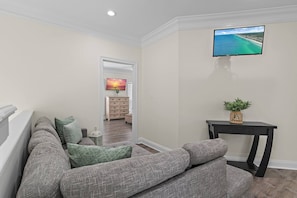 Ein Wohnzimmer mit einem grauen Sofa, einem kleinen Tisch mit einer Pflanze und einem an der Wand befestigten Fernseher.