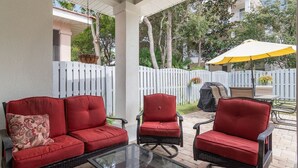 A covered patio area with red cushioned chairs and a glass-top table, surrounded by a white fence and trees. - The Cayman House: Steps from Beach | Pool & Clubhouse Amenities (Destin)