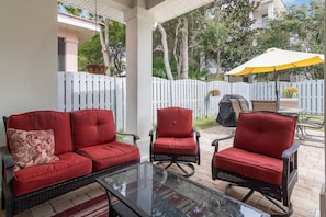 A covered patio area with red cushioned chairs and a glass-top table, surrounded by a white fence and trees. - The Cayman House: Steps from Beach | Pool & Clubhouse Amenities (Destin)