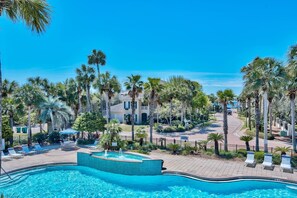 A pool area with lounge chairs and a fountain, surrounded by palm trees and a building with a tiled roof. - The Cayman House: Steps from Beach | Pool & Clubhouse Amenities (Destin)
