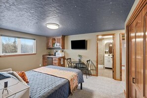 A bedroom with a bed, a desk, a chair, a television, and a kitchenette. - Nomad Lodge - 5 Bedroom Nature & Travel Inspired Home, Centrally Located (Wasilla)