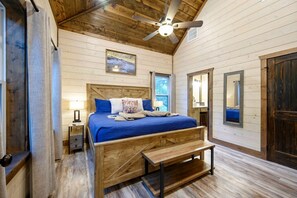 A wooden cabin bedroom with a bed, a bench, a ceiling fan, and a window. - Peaceful Easy Feeling · Woodland Retreat: Hot Tub, Swing Chairs, Fire Pit (Broken Bow)