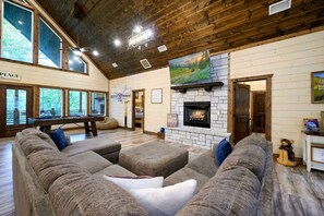 A cozy living room with a stone fireplace, large windows, and a pool table. - Peaceful Easy Feeling · Woodland Retreat: Hot Tub, Swing Chairs, Fire Pit (Broken Bow)