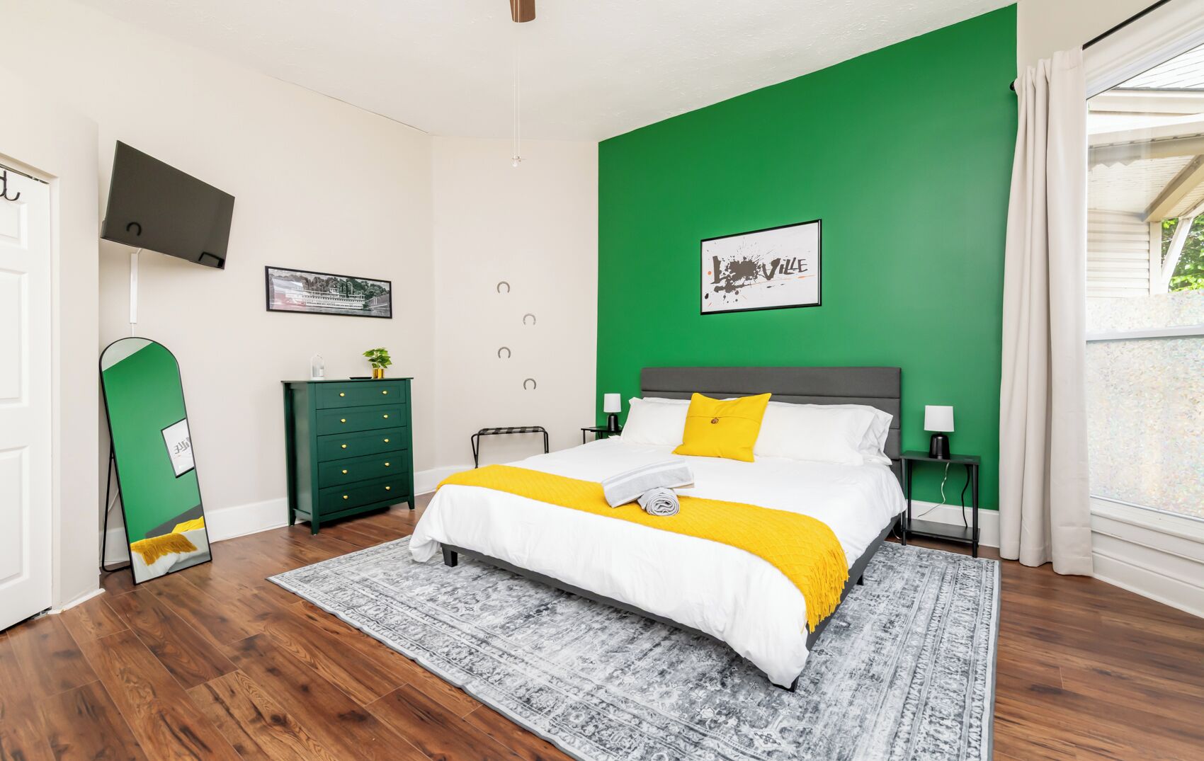 Ein Schlafzimmer mit einem Bett, einer grünen Wand, einem an der Wand befestigten Fernseher, einer grünen Kommode und einem Fenster mit Vorhängen.