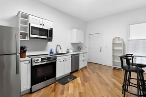 A modern kitchen with white cabinetry, a built-in microwave, a stove, and a dishwasher. There is a small dining area with a table and two chairs. - Louisville Charm 5BR Retreat | Expo | Dwntwn (Louisville)
