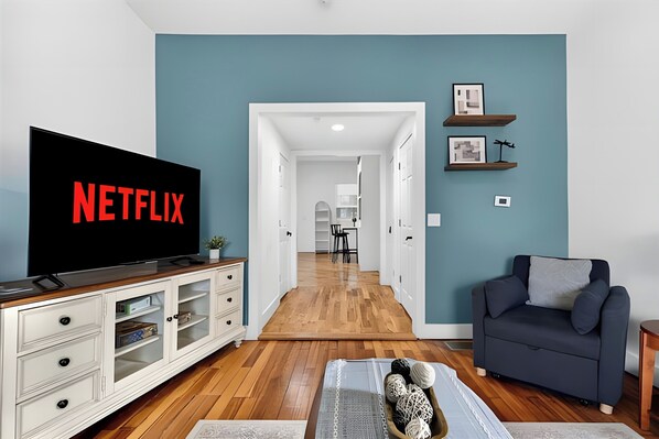 A living room with a television displaying 'NETFLIX,' a sofa, a coffee table, and a hallway leading to a kitchen.