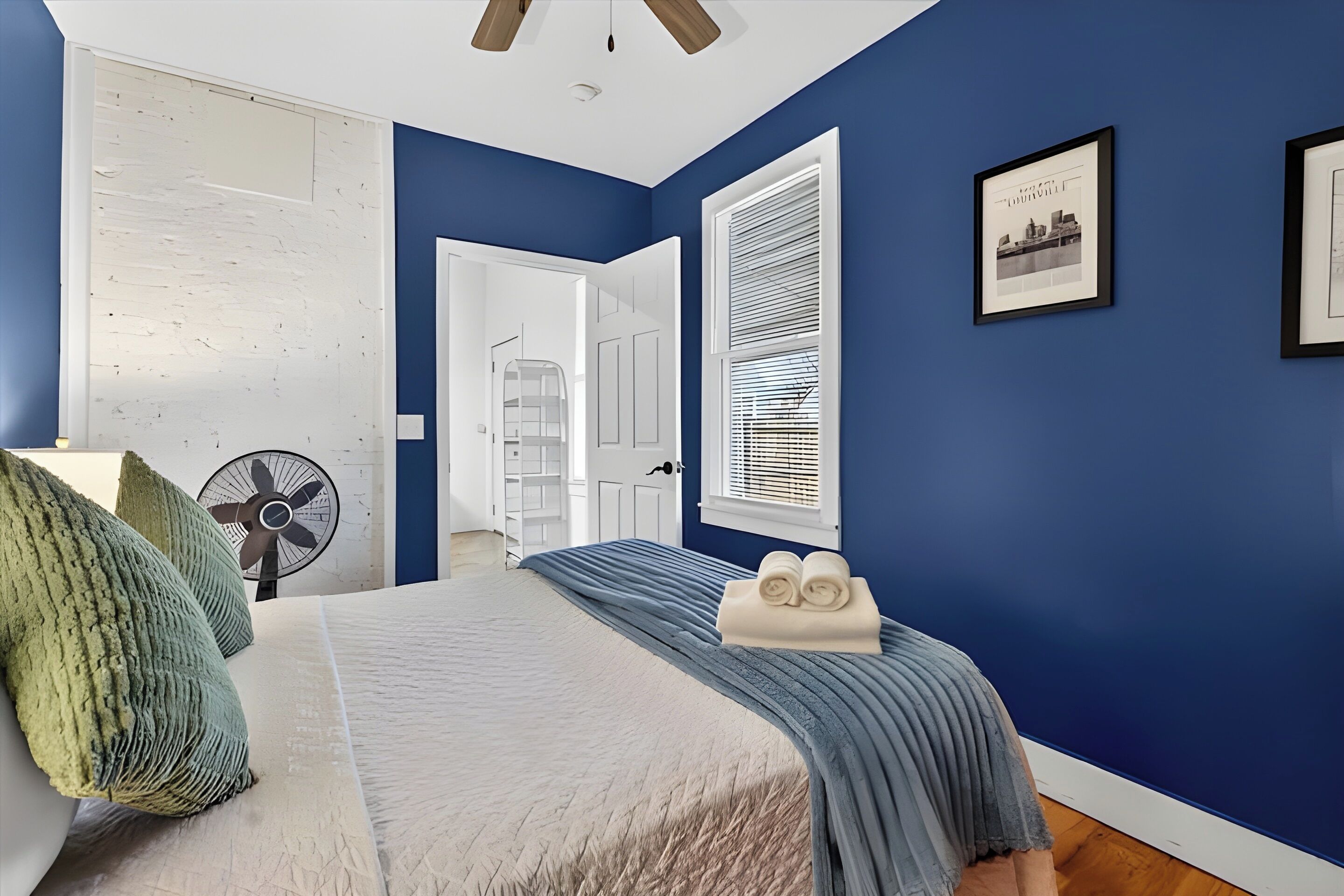 A bedroom with a bed, a fan, a window with blinds, and a door leading to another room.