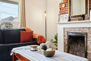 Sala de estar com sofá preto, almofadas laranja, mesa branca com uma tigela e lareira de tijolos.
